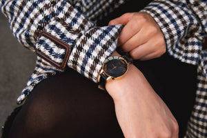 a woman wearing a brown and cream checked coat, is rolling up her sleeve to show our bold spirt watch in black and gold. 